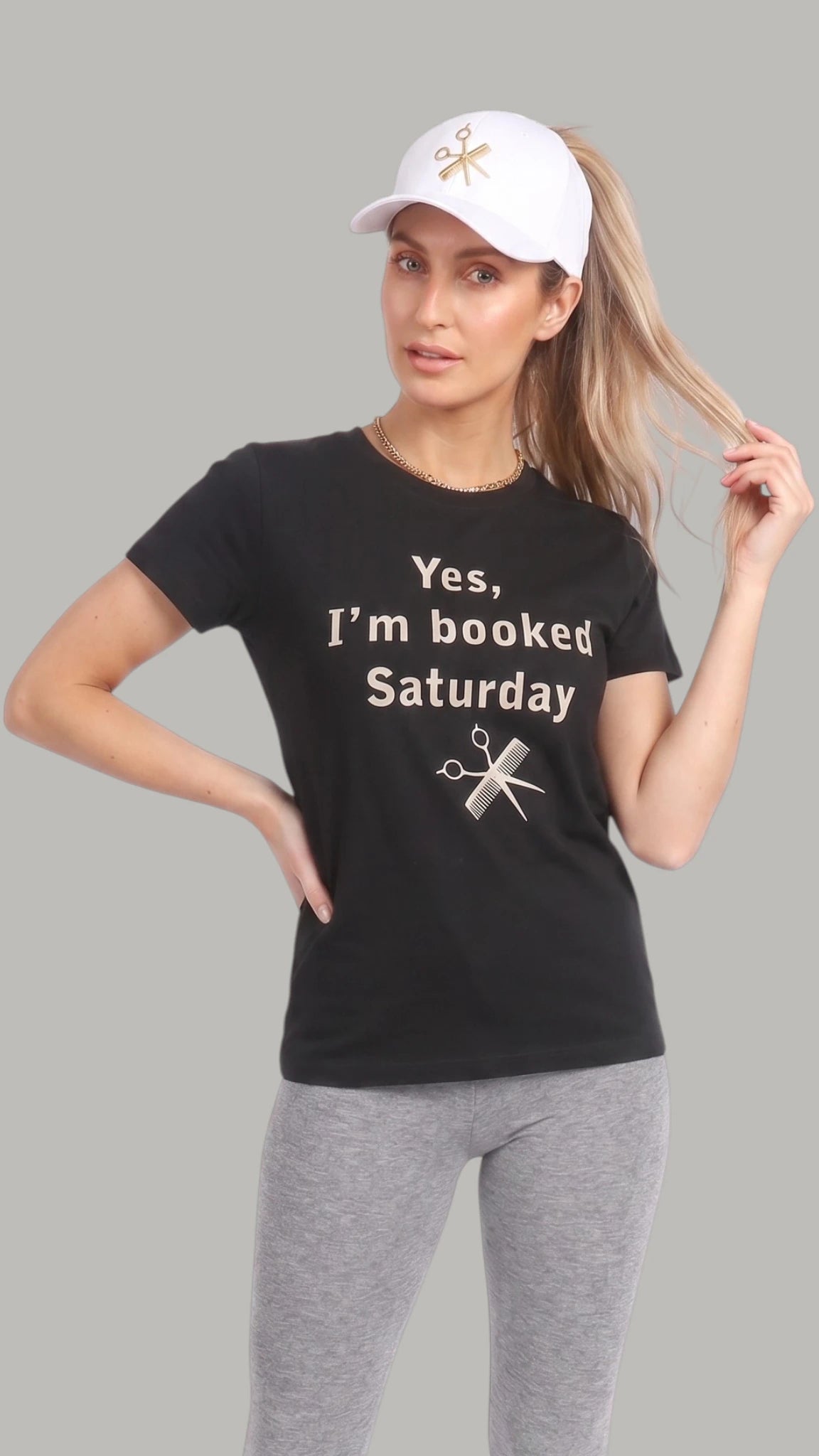 Three-quarter front view of a blonde woman wearing a white Ladybird Line ponytail baseball cap with a gold embroidered shear and comb emblem, styled with a high ponytail, black graphic t-shirt, and heather grey leggings against a solid light grey background.