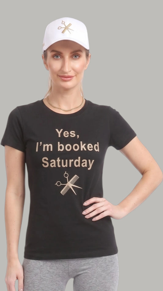 Front view of a woman with a high ponytail modeling a white Ladybird Line baseball cap with a gold embroidered shear and comb emblem, paired with a black "Yes, I'm booked Saturday" graphic t-shirt and heather grey leggings against a solid light grey background.