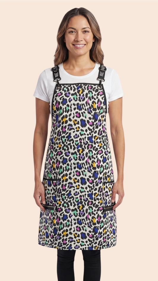 Front view of a dark-haired woman smiling while modeling the Ladybird Line 137 Multi Color Leopard Print Waterproof Signature Strap Crossover Apron, featuring a vibrant multi-color leopard print, black signature crossover straps, and zippered lapel pockets, worn over a white t-shirt and black pants against a solid pale peach background.
