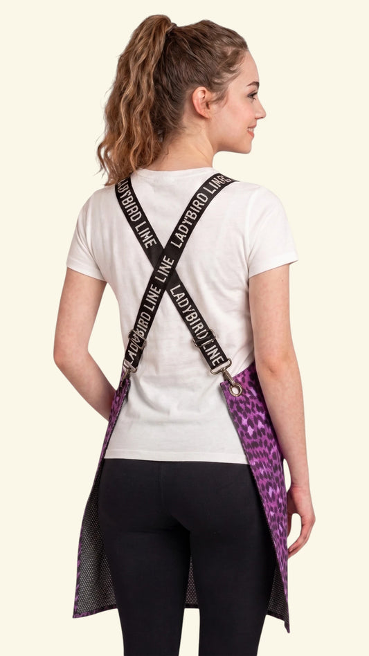 Back view of a dark-haired woman modeling the Ladybird Line 138 Pink Leopard Print Waterproof Signature Strap Crossover Apron, showing the crisscross black signature straps with white 'LADY BIRD LINE' text over a white t-shirt and black leggings against a solid cream background.