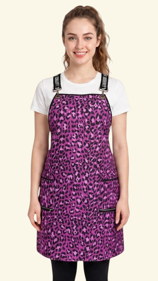 Front view of a woman with dark hair tied back in a ponytail smiling as she models the Ladybird Line 138 Pink Leopard Print Waterproof Signature Strap Crossover Apron, featuring a vibrant pink and purple leopard print, black signature crossover straps, and zippered lapel pockets, worn over a white t-shirt and black leggings against a solid cream background.