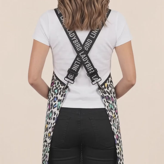 Back view of a dark-haired woman modeling the Ladybird Line 137 Multi Color Leopard Print Waterproof Signature Strap Crossover Apron, showing the crisscross black signature straps with white 'LADYBIRD LINE' text over a white t-shirt and black pants against a solid pale peach background.