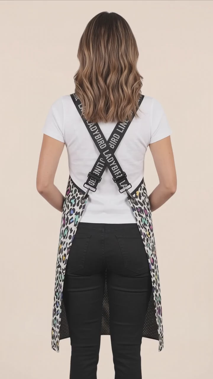 Back view of a dark-haired woman modeling the Ladybird Line 137 Multi Color Leopard Print Waterproof Signature Strap Crossover Apron, showing the crisscross black signature straps with white 'LADYBIRD LINE' text over a white t-shirt and black pants against a solid pale peach background.