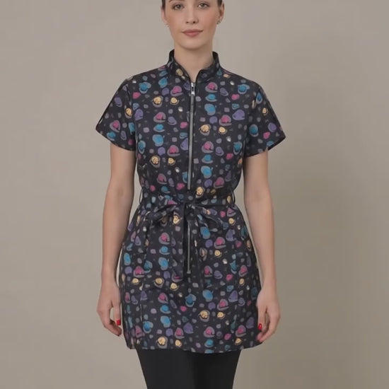 A dark-haired woman smiling while modeling the Ladybird Line 213 Rainbow Half Zipper Mini Paw Print Water Resistant Scrub Tunic, featuring a black short-sleeve tunic with an all-over colorful mini paw print pattern, rainbow half-zipper, mandarin collar, front pockets, and an adjustable waist tie belt, worn over dark pants against a solid light beige background.