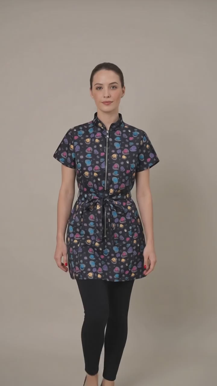 A dark-haired woman smiling while modeling the Ladybird Line 213 Rainbow Half Zipper Mini Paw Print Water Resistant Scrub Tunic, featuring a black short-sleeve tunic with an all-over colorful mini paw print pattern, rainbow half-zipper, mandarin collar, front pockets, and an adjustable waist tie belt, worn over dark pants against a solid light beige background.