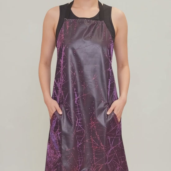 An Asian woman smiling while modeling the Ladybird Line 141 Rose Sparkle Waterproof & Bleach Resistant Apron in motion, swaying side to side with hands in her pockets over a black top and leggings against a light gray background.