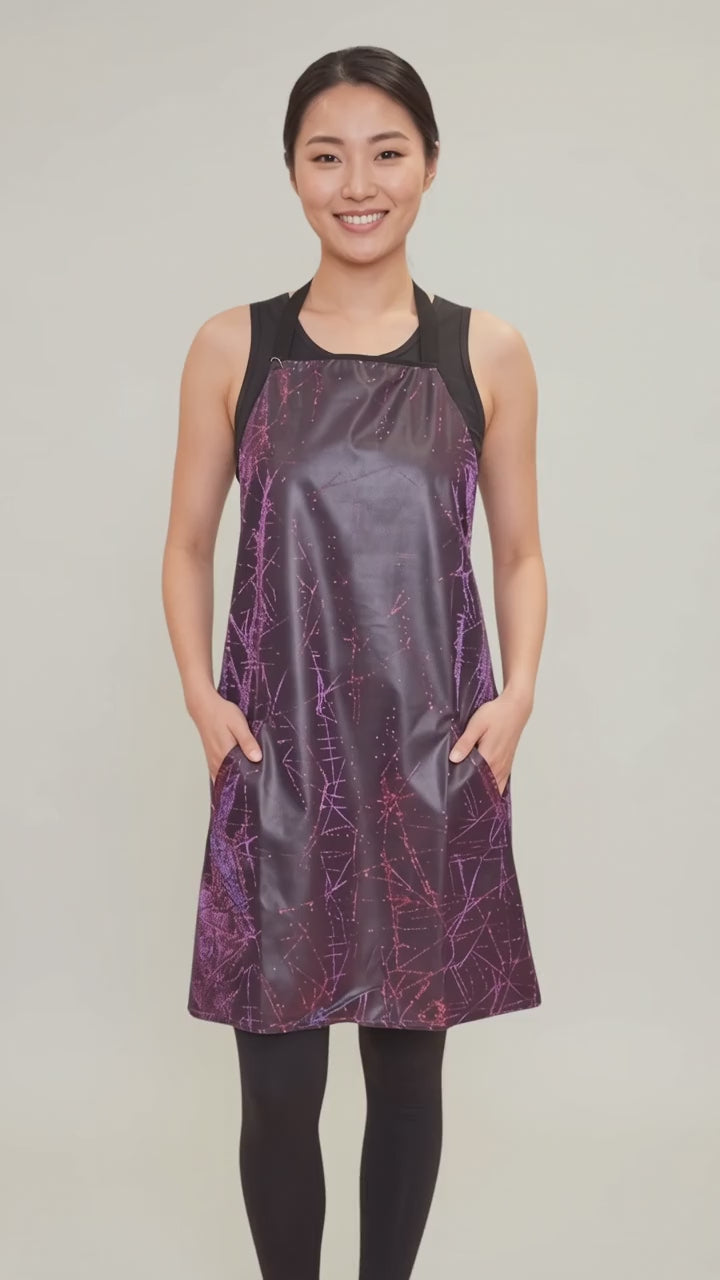 An Asian woman smiling while modeling the Ladybird Line 141 Rose Sparkle Waterproof & Bleach Resistant Apron in motion, swaying side to side with hands in her pockets over a black top and leggings against a light gray background.