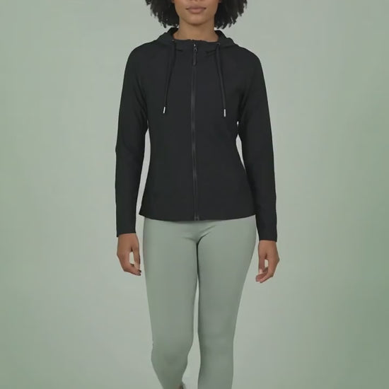 A woman with short curly dark hair modeling the Ladybird Line 122 Black Shadow Hooded Hair & Bleach Resistant Grooming Jacket in motion, walking forward zipped up over light gray leggings and athletic shoes against a light green background.