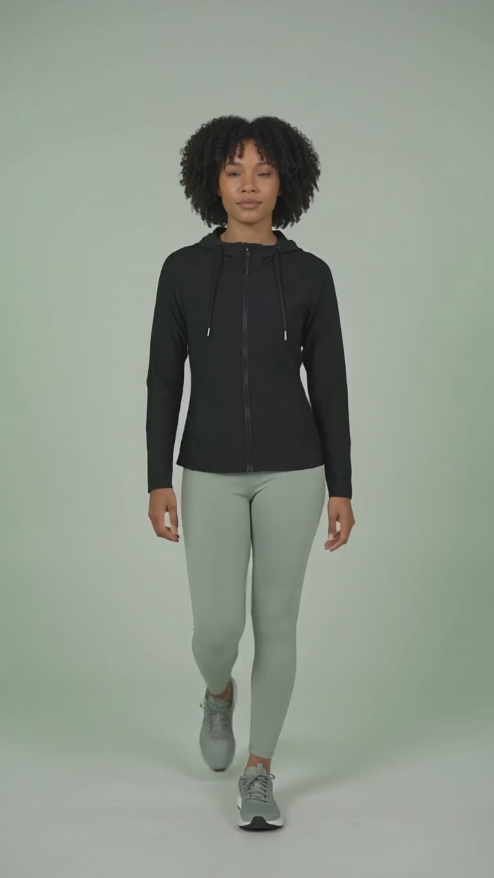 A woman with short curly dark hair modeling the Ladybird Line 122 Black Shadow Hooded Hair & Bleach Resistant Grooming Jacket in motion, walking forward zipped up over light gray leggings and athletic shoes against a light green background.