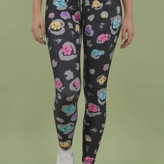 A woman modeling the Ladybird Line 126 Paw Print Shadow Hair & Bleach Resistant Grooming Leggings in motion, featuring a colorful pink, teal, purple, and yellow paw print pattern, worn with a black top and white sneakers against a light green background.
