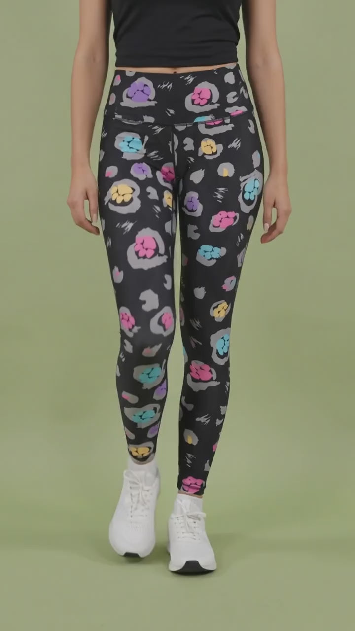 A woman modeling the Ladybird Line 126 Paw Print Shadow Hair & Bleach Resistant Grooming Leggings in motion, featuring a colorful pink, teal, purple, and yellow paw print pattern, worn with a black top and white sneakers against a light green background.