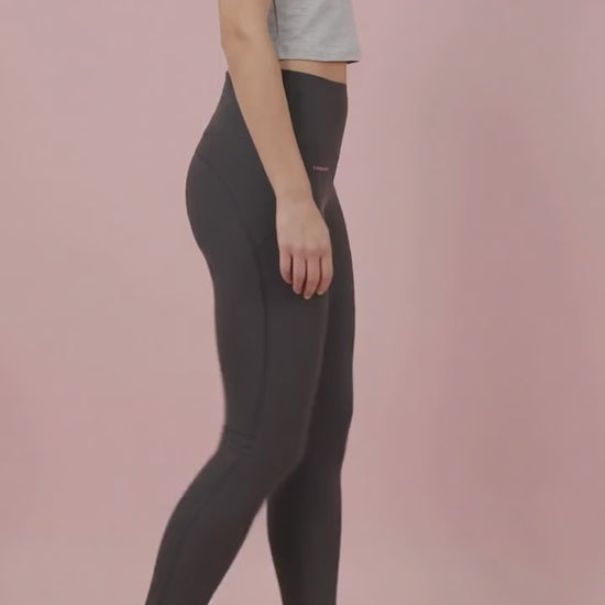 A woman modeling the solid black Ladybird Line 125 Black Shadow Hair & Bleach Resistant Grooming Leggings in motion, walking in profile wearing a gray cropped t-shirt and white sneakers against a pale pink background.