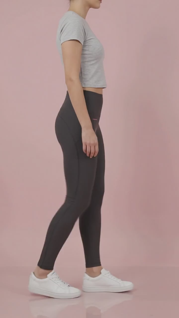 A woman modeling the solid black Ladybird Line 125 Black Shadow Hair & Bleach Resistant Grooming Leggings in motion, walking in profile wearing a gray cropped t-shirt and white sneakers against a pale pink background.