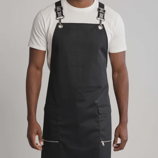 A man smiling while modeling the Ladybird Line 667 Waterproof Black Signature Strap Crossover Apron, featuring a solid black apron with black signature crossover straps with white text, zippered lapel pockets, worn over a white t-shirt and blue jeans against a solid off-white background.