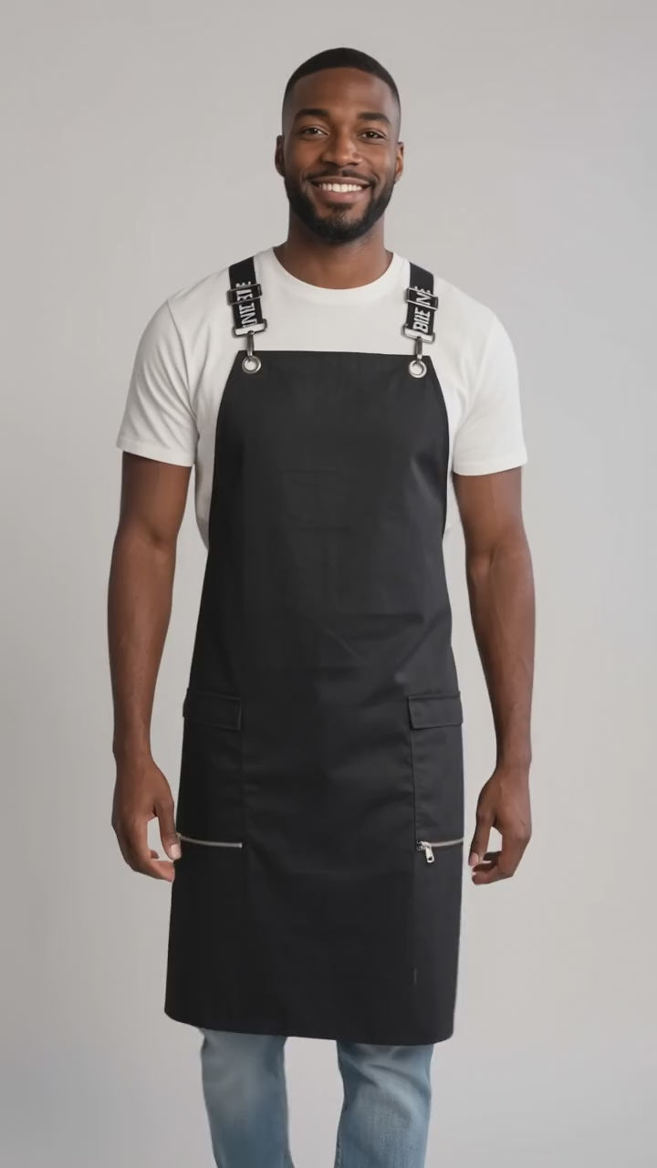 A man smiling while modeling the Ladybird Line 667 Waterproof Black Signature Strap Crossover Apron, featuring a solid black apron with black signature crossover straps with white text, zippered lapel pockets, worn over a white t-shirt and blue jeans against a solid off-white background.