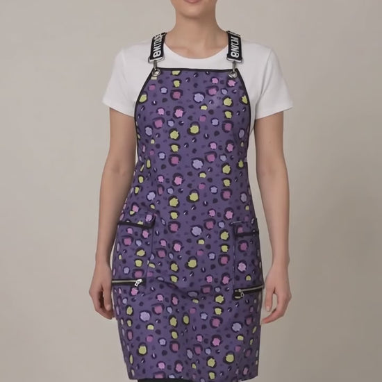 A woman modeling the Ladybird Line 139 Lilac Mini Paw Print Waterproof Signature Strap Crossover Apron in motion, swaying side to side over a white t-shirt and black pants against a light beige background.