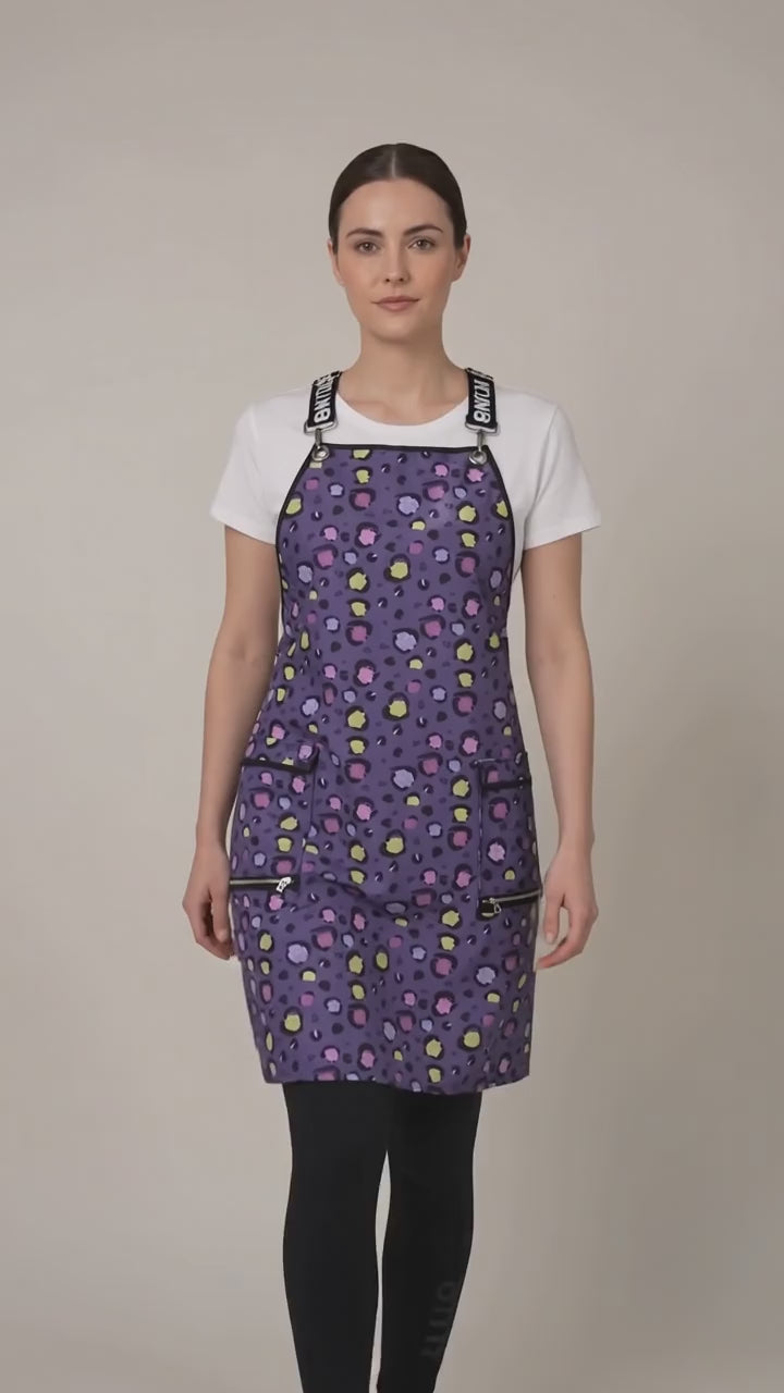 A woman modeling the Ladybird Line 139 Lilac Mini Paw Print Waterproof Signature Strap Crossover Apron in motion, swaying side to side over a white t-shirt and black pants against a light beige background.