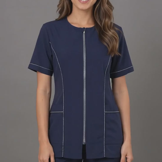 A brunette woman smiling while modeling the Ladybird Line 854 Navy Rhinestone Trim Waterproof Grooming Smock, featuring a navy short-sleeve zip-up smock with rhinestone trim along the zipper, sleeves, and pockets, paired with matching navy pants against a neutral grey background.