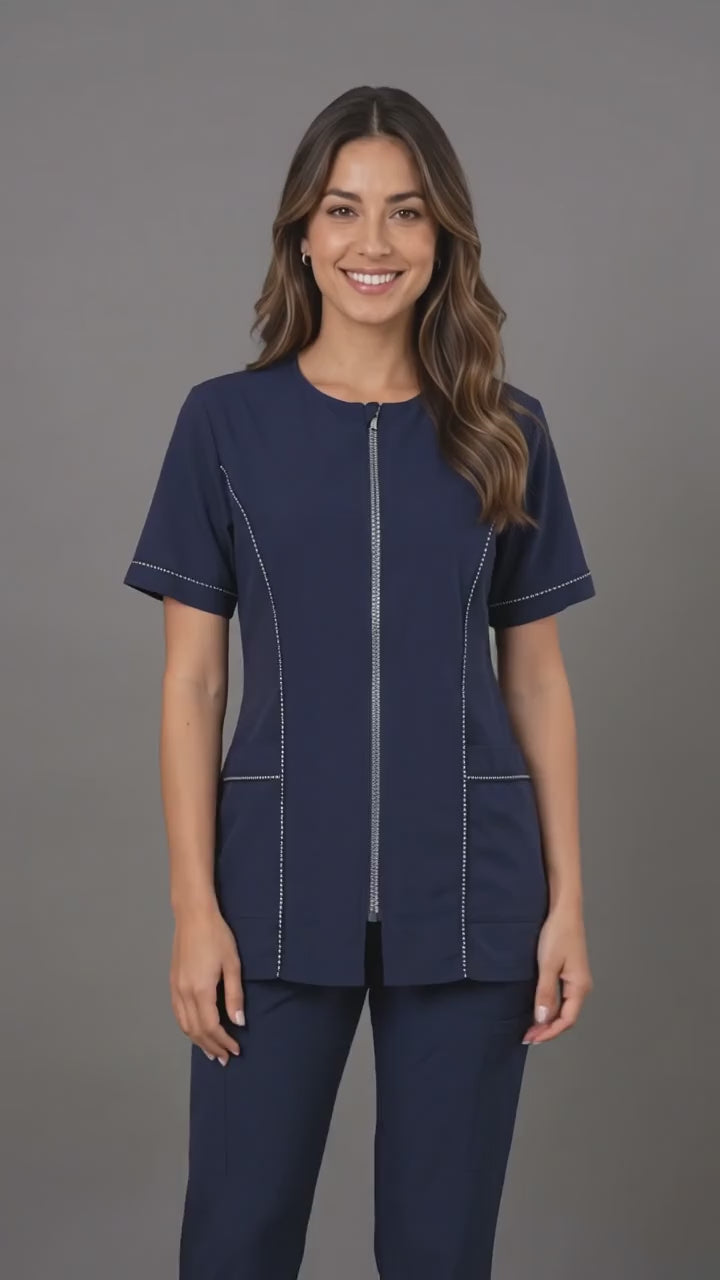 A brunette woman smiling while modeling the Ladybird Line 854 Navy Rhinestone Trim Waterproof Grooming Smock, featuring a navy short-sleeve zip-up smock with rhinestone trim along the zipper, sleeves, and pockets, paired with matching navy pants against a neutral grey background.