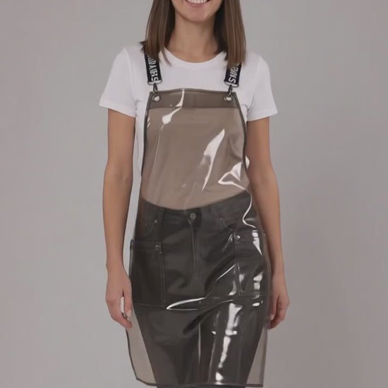 A dark-haired woman smiling while modeling the Ladybird Line 113 Waterproof Black Clear Crossover Apron, featuring a clear waterproof plastic body, black signature crossover straps with white text, and front pockets, worn over a white t-shirt and dark pants against a solid light grey background.