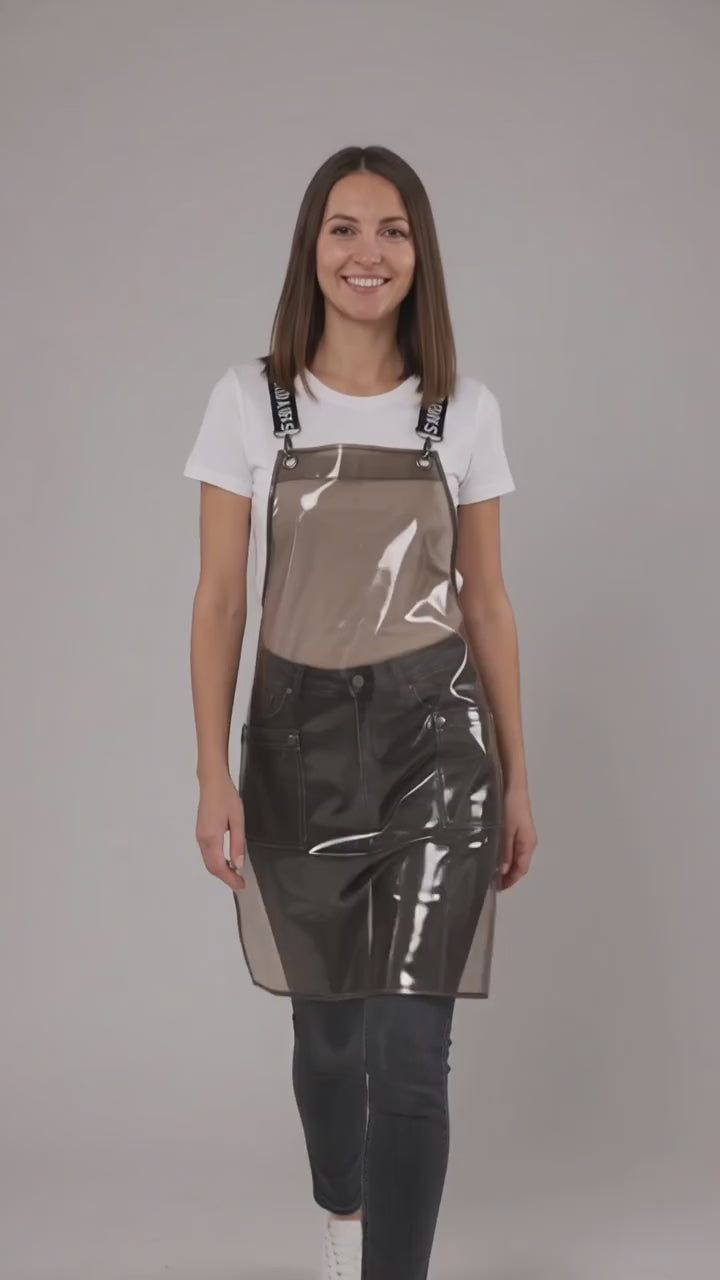 A dark-haired woman smiling while modeling the Ladybird Line 113 Waterproof Black Clear Crossover Apron, featuring a clear waterproof plastic body, black signature crossover straps with white text, and front pockets, worn over a white t-shirt and dark pants against a solid light grey background.