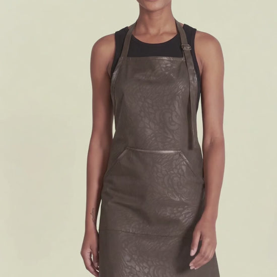 A woman with short blonde hair modeling the Ladybird Line 130 Black Leopard Print Hair & Bleach Resistant Grooming Apron in motion, standing with hands at her sides over a black top against a pale yellow background.