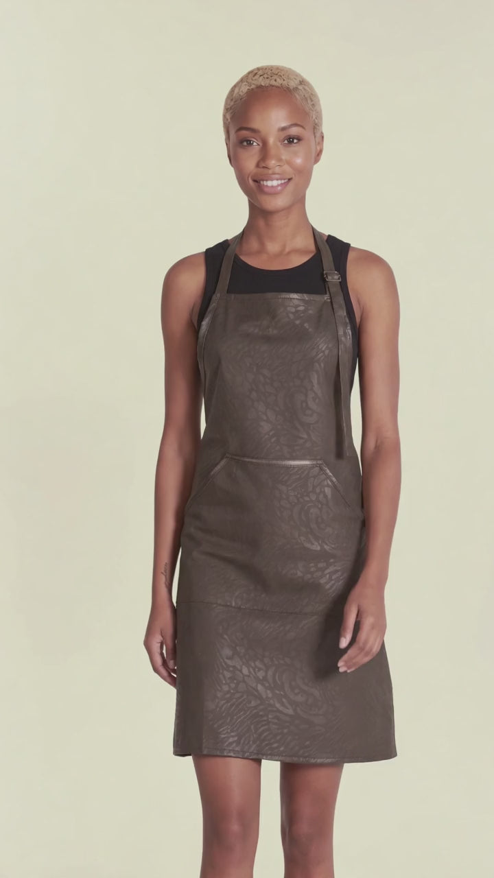 A woman with short blonde hair modeling the Ladybird Line 130 Black Leopard Print Hair & Bleach Resistant Grooming Apron in motion, standing with hands at her sides over a black top against a pale yellow background.