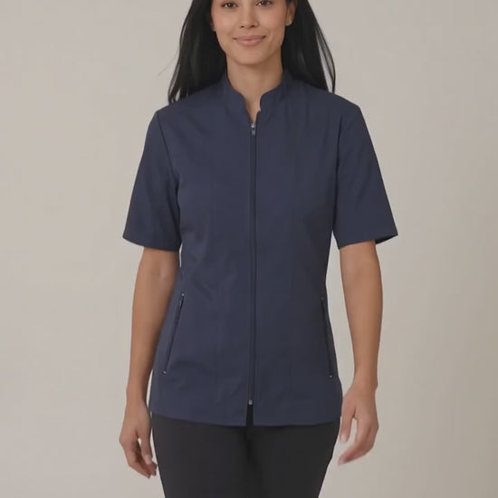 A woman smiling while modeling the Ladybird Line 427 Water Resistant Navy Mandarin Collar Tailored Barber & Grooming Jacket, featuring a navy zip-up short-sleeve jacket with a mandarin collar, paired with black pants against a solid off-white background.