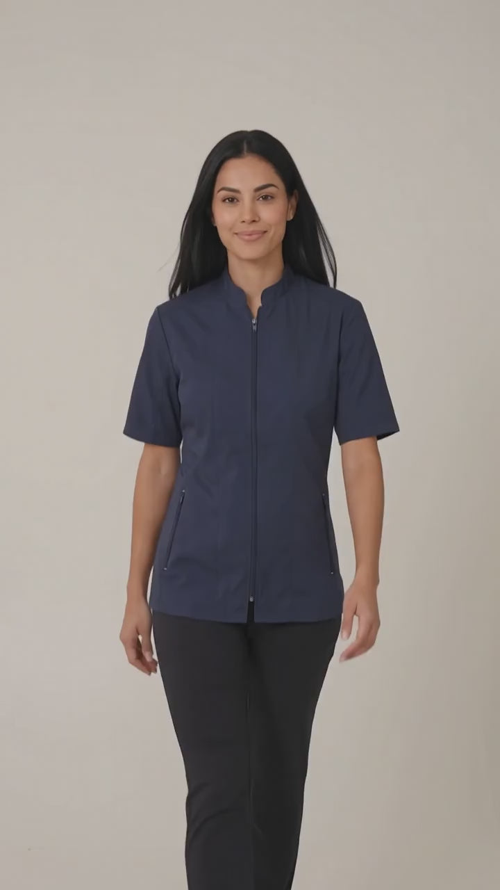A woman smiling while modeling the Ladybird Line 427 Water Resistant Navy Mandarin Collar Tailored Barber & Grooming Jacket, featuring a navy zip-up short-sleeve jacket with a mandarin collar, paired with black pants against a solid off-white background.