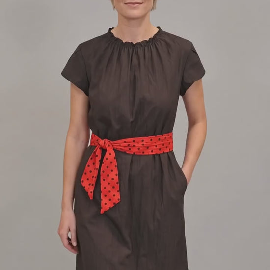 A blonde woman modeling the Ladybird Line 444 Classic Black Water Resistant Scarf Belt Professional Dress, featuring a black short-sleeve dress with a gathered neckline, elastic waist, side pockets, and a red polka dot scarf belt, worn over dark pants against a solid light grey background.