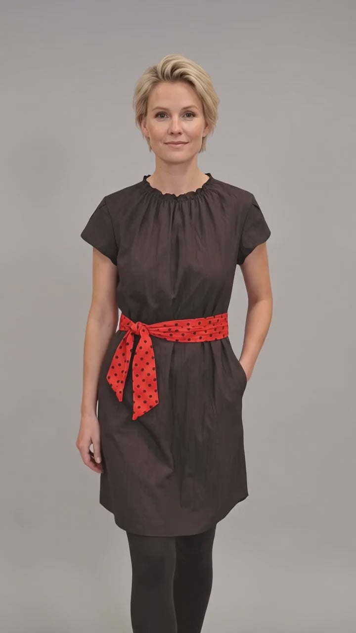 A blonde woman modeling the Ladybird Line 444 Classic Black Water Resistant Scarf Belt Professional Dress, featuring a black short-sleeve dress with a gathered neckline, elastic waist, side pockets, and a red polka dot scarf belt, worn over dark pants against a solid light grey background.