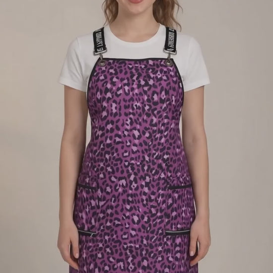 A dark-haired woman smiling while modeling the Ladybird Line 138 Pink Leopard Print Waterproof Signature Strap Crossover Apron, featuring a vibrant pink and purple leopard print, black signature crossover straps, and zippered lapel pockets, worn over a white t-shirt and black leggings against a solid cream background.