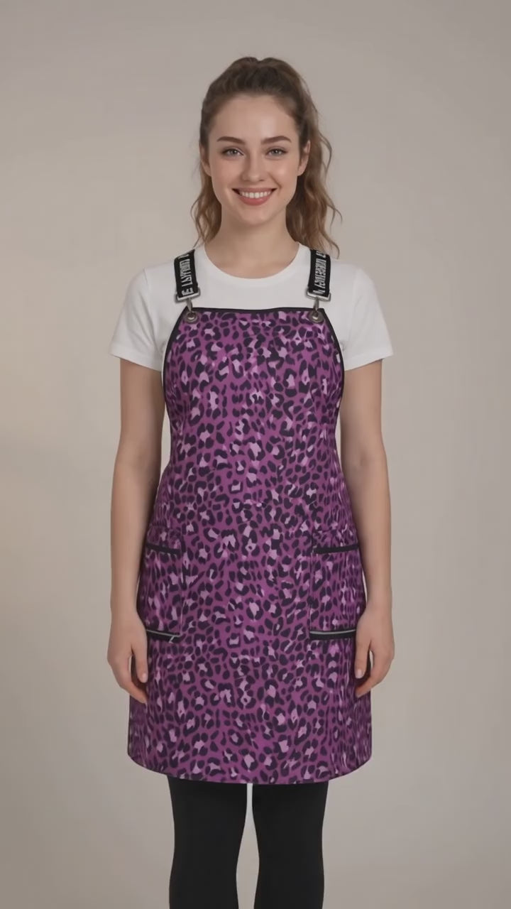 A dark-haired woman smiling while modeling the Ladybird Line 138 Pink Leopard Print Waterproof Signature Strap Crossover Apron, featuring a vibrant pink and purple leopard print, black signature crossover straps, and zippered lapel pockets, worn over a white t-shirt and black leggings against a solid cream background.