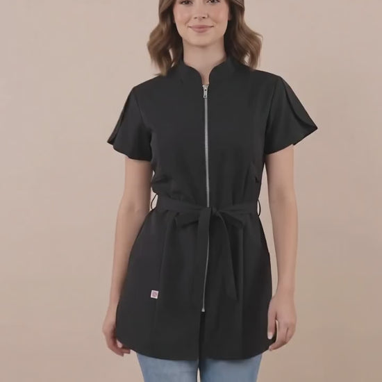 A brown-haired woman smiling while modeling the Ladybird Line 214 Classic Black Rhinestone Half Zipper Water Resistant Scrub Tunic, featuring a black short-sleeve tunic with a rhinestone half-zipper front, mandarin collar, front pockets, and an adjustable waist tie belt, worn over blue jeans against a solid light beige background.