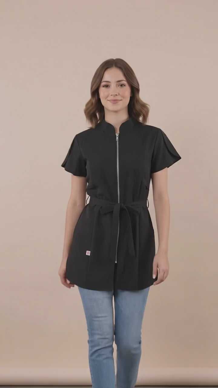 A brown-haired woman smiling while modeling the Ladybird Line 214 Classic Black Rhinestone Half Zipper Water Resistant Scrub Tunic, featuring a black short-sleeve tunic with a rhinestone half-zipper front, mandarin collar, front pockets, and an adjustable waist tie belt, worn over blue jeans against a solid light beige background.