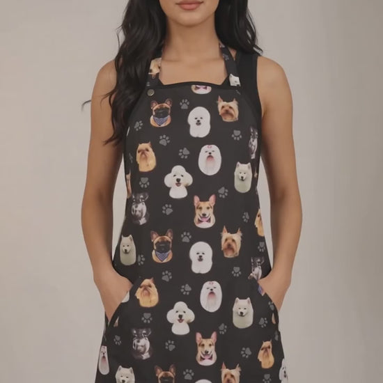 A dark-haired woman modeling the Ladybird Line 610 Dog Breeds Print Waterproof Hair & Bleach Resistant Grooming Apron, featuring a black apron with an all-over colorful dog breed head pattern, front pockets, and adjustable straps, set against a solid off-white background.