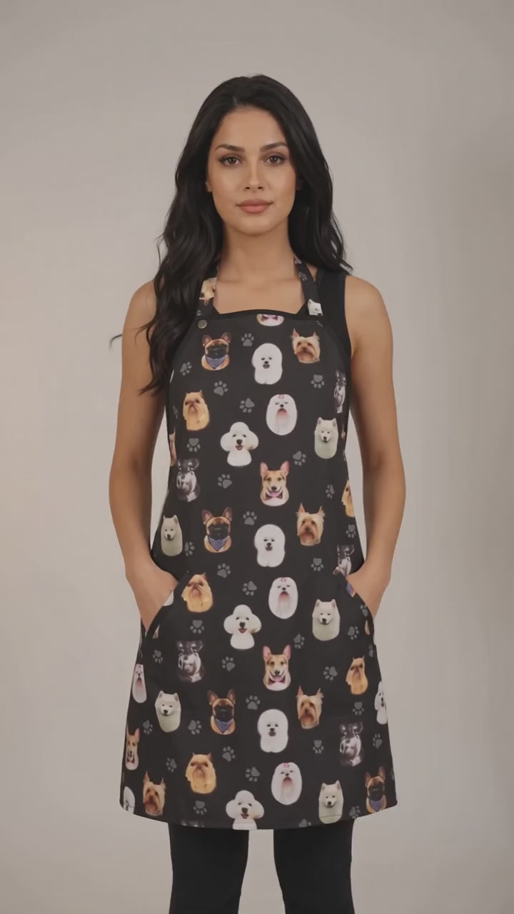 A dark-haired woman modeling the Ladybird Line 610 Dog Breeds Print Waterproof Hair & Bleach Resistant Grooming Apron, featuring a black apron with an all-over colorful dog breed head pattern, front pockets, and adjustable straps, set against a solid off-white background.