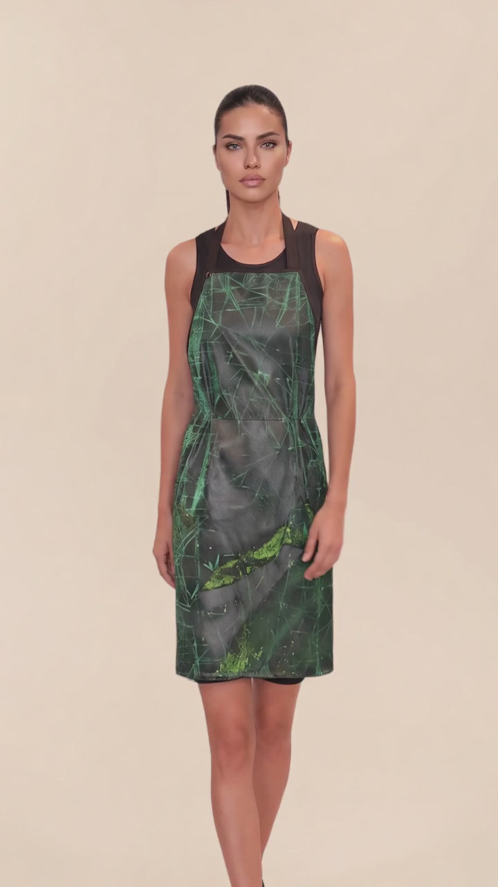 A dark-haired woman modeling the Ladybird Line 142 Green Sparkle Waterproof & Bleach Resistant Apron in motion, swaying side to side over a black top against a pale beige background.
