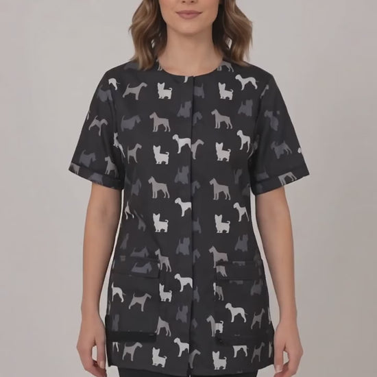 A dark-haired woman smiling while modeling the Ladybird Line 709 Grey Terrier Unisex Waterproof Grooming Smock, featuring an all-over grey, black, and white dog silhouette print with covered zipper lapels and front pockets, worn over black pants against a solid light grey background.