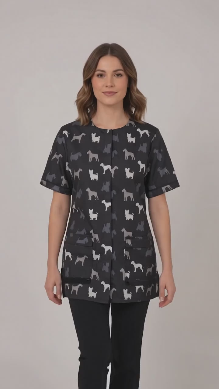 A dark-haired woman smiling while modeling the Ladybird Line 709 Grey Terrier Unisex Waterproof Grooming Smock, featuring an all-over grey, black, and white dog silhouette print with covered zipper lapels and front pockets, worn over black pants against a solid light grey background.