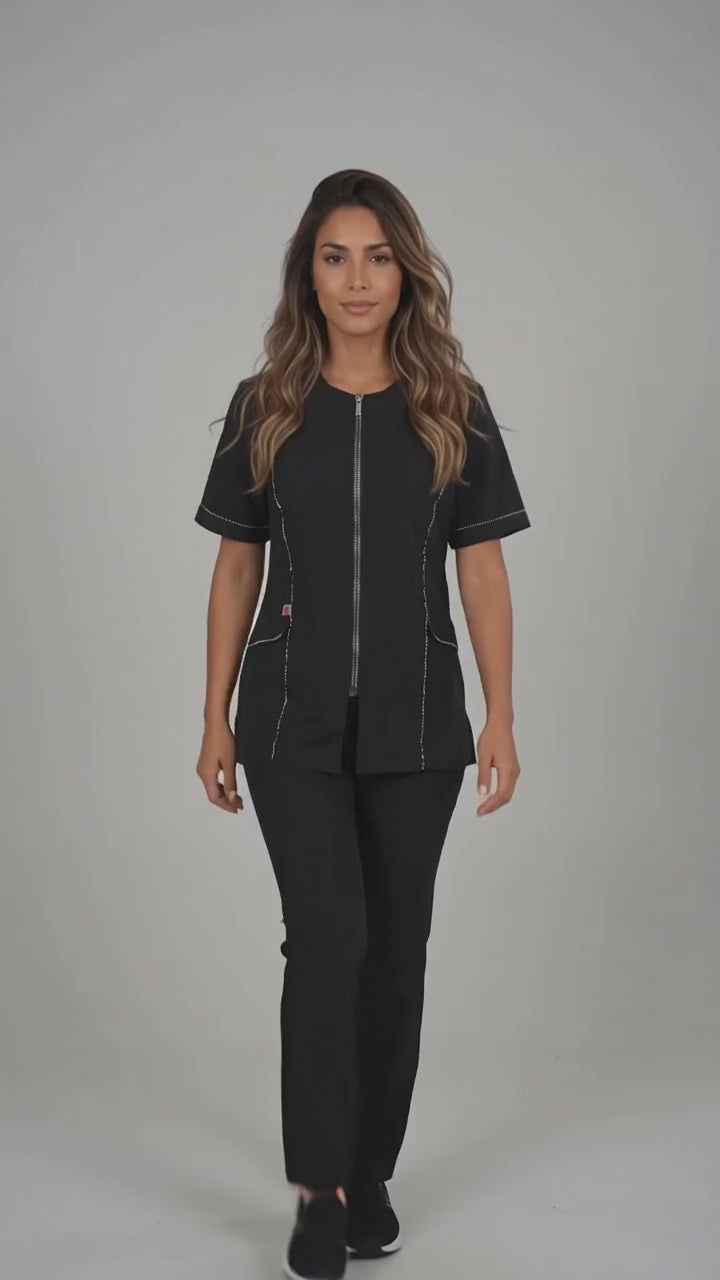 A brown-haired woman smiling while modeling the Ladybird Line 850 Rhinestone Trim Water, Bleach & Hair Resistant Styling Jacket, featuring a black short-sleeve styling jacket with sparkling rhinestone trim along the zipper and front lapel, worn with black pants against a solid light grey background.