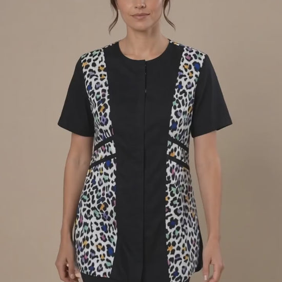 A dark-haired woman smiling while modeling the Ladybird Line 737 Multi Color Leopard Color Block Waterproof Grooming Smock, featuring a black center panel with vibrant multi-color leopard print side panels, worn over black pants against a solid beige background.