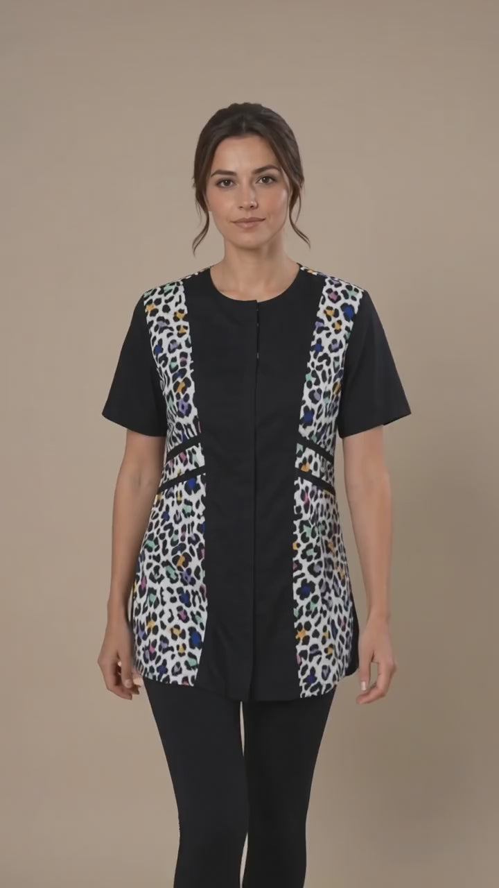 A dark-haired woman smiling while modeling the Ladybird Line 737 Multi Color Leopard Color Block Waterproof Grooming Smock, featuring a black center panel with vibrant multi-color leopard print side panels, worn over black pants against a solid beige background.